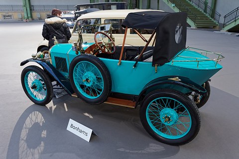 Side view of a turquoise car with a pointed tail, luggage rack and black hood, a visitor walking behind. Opens a larger view