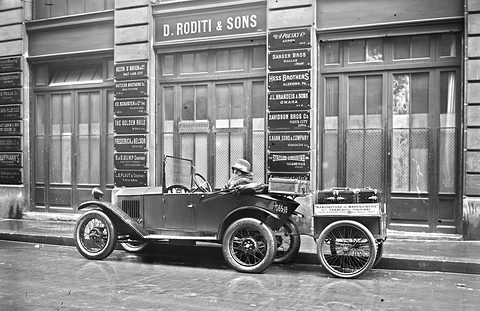 A small open car with a two-wheeled box trailer at the kerb before an office front hung with company name plates.