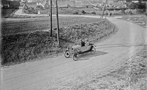 A small open two-seater with the number 45 on its bonnet rounding a sweeping dirt road, a village and telegraph poles beyond. Opens a larger view