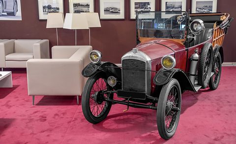 A red open two-seater with a nickel radiator and wire wheels on red carpet beside a cream sofa and framed photographs. Opens a larger view