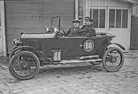 Two men in caps seated side by side in a small open tourer with wire wheels and a roundel marked 66, before a shuttered garage. Opens a larger view