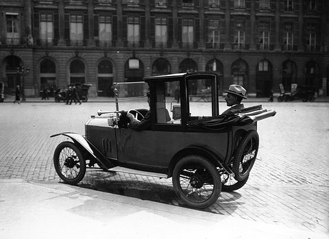 A small taxi with a tall enclosed cab at the back and an open driver’s seat in front, on cobbles before a long arcaded façade. Opens a larger view