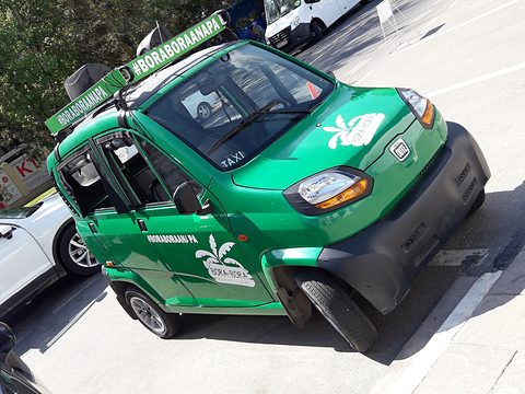High, tilted view of a green tiny four-door taxi with a roof sign and palm graphics, parked on tarmac. Opens a larger view