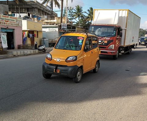 Yellow tiny four-door car driving along a city road with a red lorry behind and shops at the side.