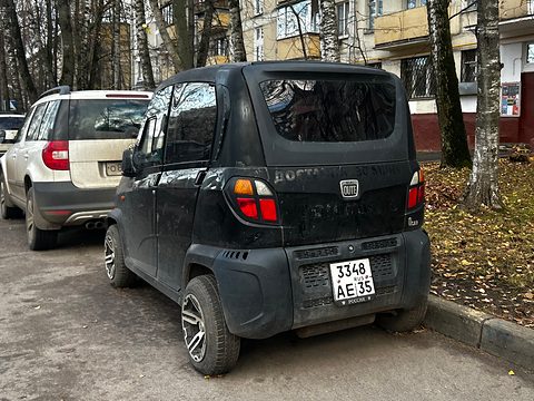 Rear of a dusty black tiny four-door car with a Russian plate, parked under birch trees by a block of flats. Opens a larger view