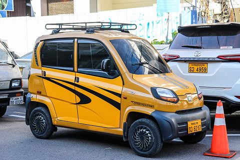 Yellow tiny four-door car with black swoosh graphics and a roof rack, parked beside a white SUV and a traffic cone. Opens a larger view