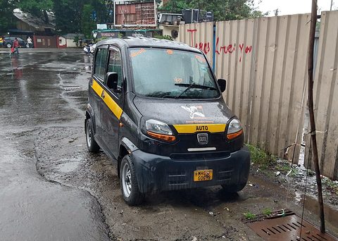 Black tiny four-door car with a yellow stripe and AUTO lettering parked on a wet street by a metal fence.
