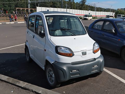 White tall tiny four-door car with a grey bumper and orange-trimmed lamps, parked beside a blue saloon.