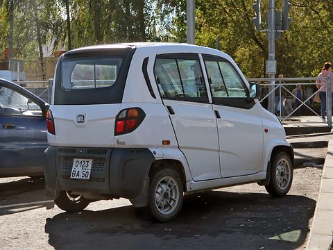 Rear of a white tiny four-door car with a Russian plate and round tail lamps, parked by a fence and trees. Opens a larger view