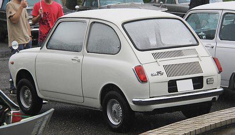 Rear three-quarter of a white small saloon with a louvred engine lid, visitors standing behind. Opens a larger view