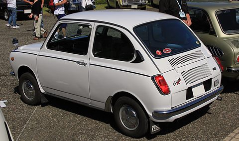 Rear three-quarter of a white small saloon with a black-and-chrome louvred tail panel, people behind. Opens a larger view