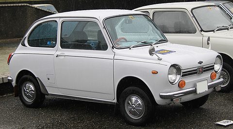 White small saloon from the front side, a slim chrome grille between round headlamps, other cars behind. Opens a larger view
