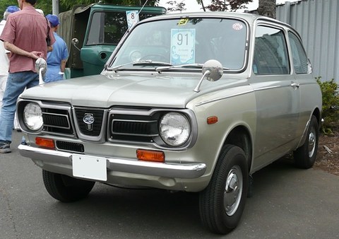Grey small saloon with a black slatted grille between round headlamps and amber indicators, a man standing at left. Opens a larger view