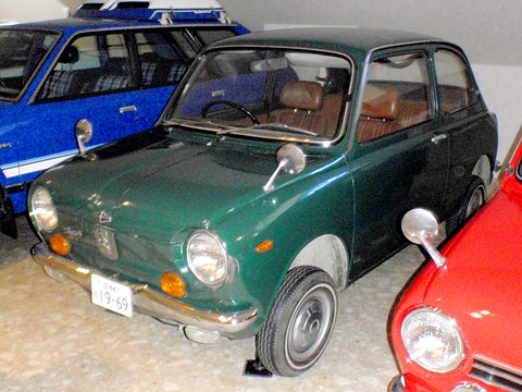 Dark green small saloon between a blue car and a red car on a beige museum floor. Opens a larger view