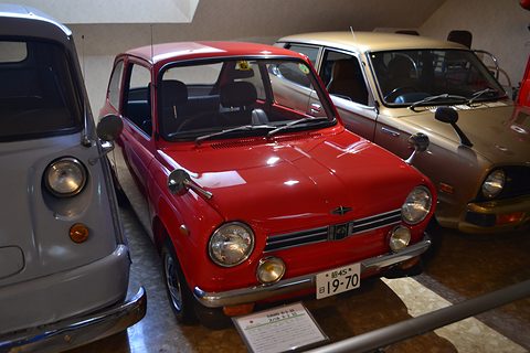 Red small saloon from the front right in a museum row, a grey van at left and a bronze car behind. Opens a larger view
