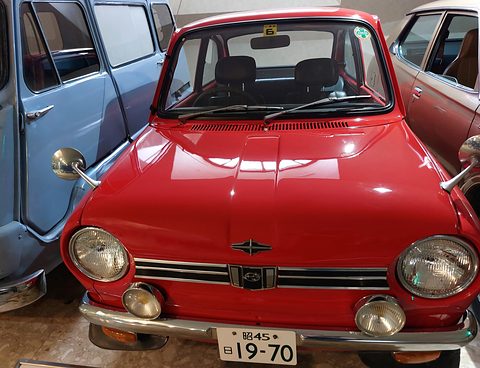 Red small saloon seen from the front, round headlamps, twin fog lamps and a number plate, between other cars.
