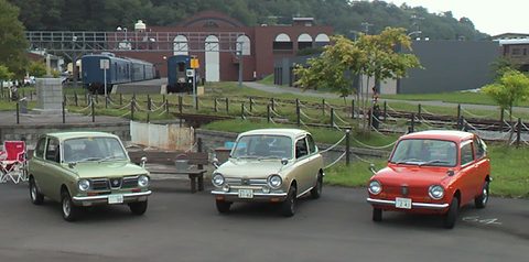 Green, cream and red small saloons parked in a row in front of a railway museum with old carriages. Opens a larger view