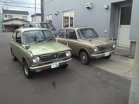 Two small saloons, one olive green and one bronze, parked on tarmac beside a grey metal building.