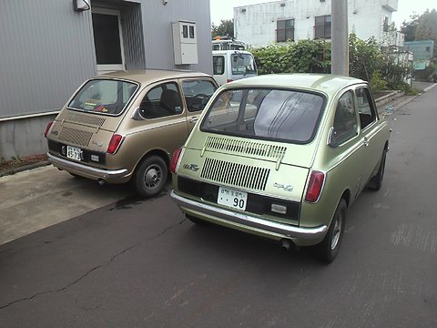 Rear view of two small saloons, green and bronze, with louvred tails, parked by a grey building. Opens a larger view