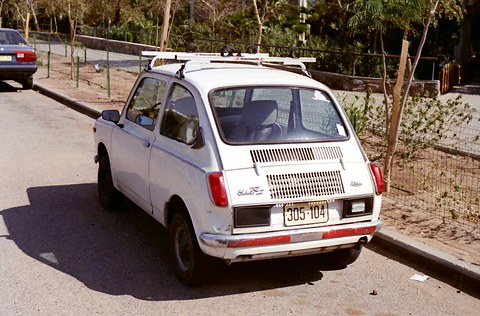 Rear three-quarter of a white small saloon with a roof rack and rows of cooling slots in the engine lid. Opens a larger view
