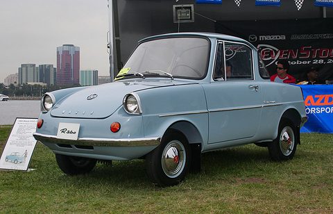 A small pale blue two-door coupé with a pointed nose, round headlamps and MAZDA letters, a show placard beside it. Opens a larger view