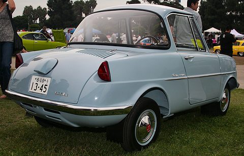 Rear three-quarter view of a small pale blue coupé with a louvred engine lid, round fuel filler and chrome bumper. Opens a larger view