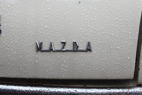 Chrome spaced letters reading MAZDA on a cream panel covered with raindrops. Opens a larger view