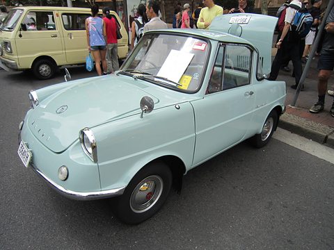 Pale blue small coupé with a sheet of paper on its windscreen, visitors crowding behind. Opens a larger view
