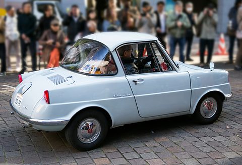 Pale grey-blue small coupé on stone paving with a crowd of spectators behind. Opens a larger view