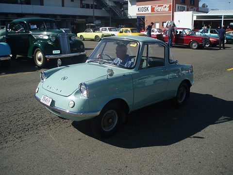 Pale blue-green small coupé on tarmac with old cars and a kiosk behind. Opens a larger view