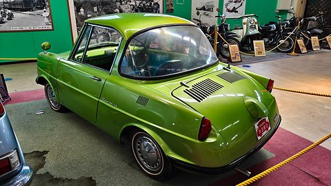 Rear three-quarter of a lime-green coupé inside a yellow rope, motorcycles behind. Opens a larger view