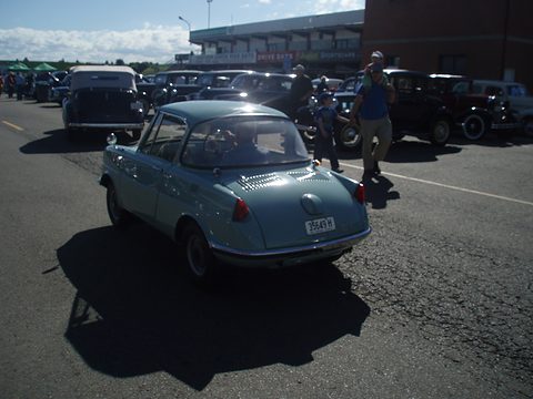 A small pale blue coupé seen from behind crossing a paddock, cars and people beyond. Opens a larger view