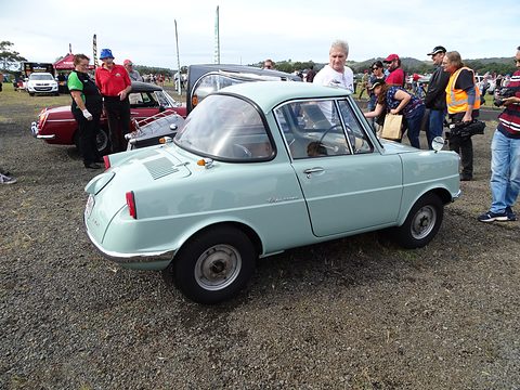 Side view of a pale green coupé on gravel, onlookers standing behind. Opens a larger view