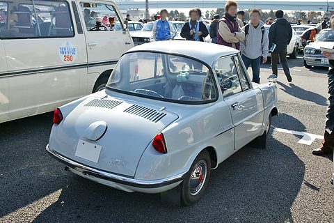 Rear three-quarter of a silver small coupé with a louvred engine lid, visitors and a white van behind. Opens a larger view