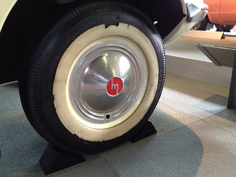 Close view of a whitewall tyre and a chrome hub cap with a red centre and an m emblem. Opens a larger view