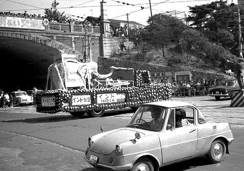 Black-and-white street scene: a small coupé with two occupants in front of a flower-covered float lettered INDIA.