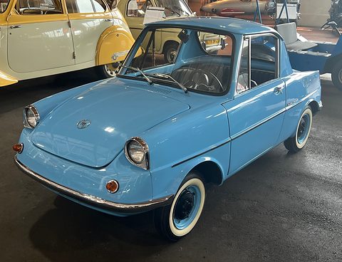Light blue small coupé on a dark floor with whitewall tyres, a yellow car behind. Opens a larger view