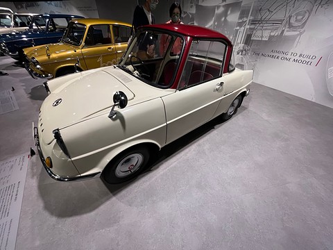 Cream small coupé with a maroon roof on a grey floor in front of a graphic wall. Opens a larger view