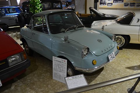 Pale blue small coupé behind a rail with placards on the floor, a white car behind. Opens a larger view