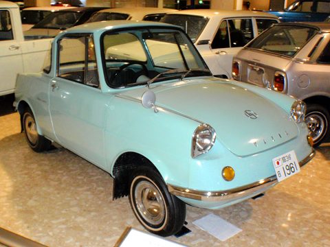 Pale turquoise small coupé on a beige museum floor among other cars. Opens a larger view