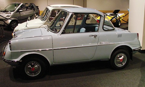 Side view of a grey small coupé on a museum floor, other small cars behind. Opens a larger view