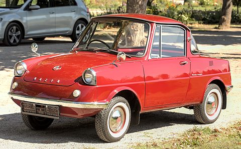 Maroon coupé seen low from the front left on gravel, a modern car behind. Opens a larger view