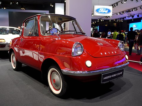 Red small coupé with whitewall tyres on a dark show stand under bright lights. Opens a larger view