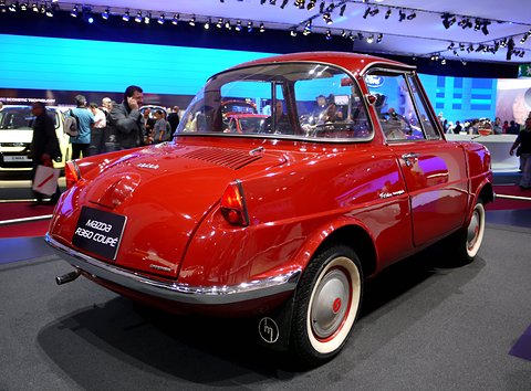 Rear three-quarter of a red coupé on a stand, a blue-lit screen and visitors behind. Opens a larger view