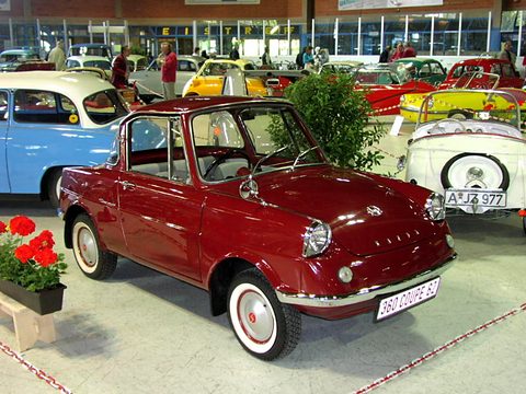 Red small coupé with whitewall tyres on a show stand edged with flowers, other old cars behind. Opens a larger view