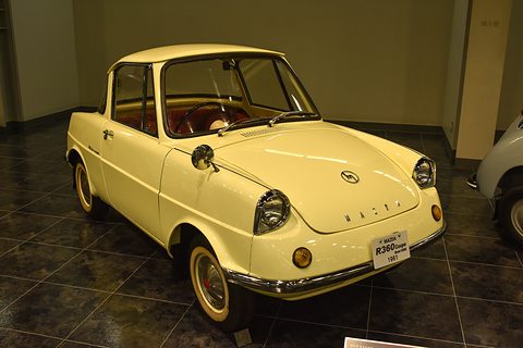 Cream small coupé with a red interior on a dark tiled floor, a museum placard on its bumper. Opens a larger view