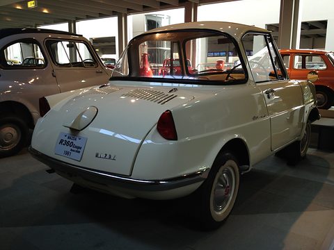 Rear three-quarter of a cream coupé with a louvred lid and a placard on its bumper. Opens a larger view