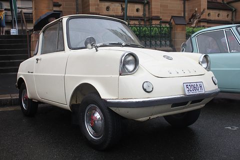 Cream small coupé with an NSW historic vehicle plate, parked on wet tarmac. Opens a larger view