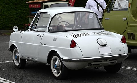 Rear three-quarter of a white coupé with a louvred tail, parked on tarmac with other cars. Opens a larger view