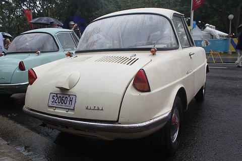 Rear three-quarter of a cream coupé with a louvred lid and a round filler cap, umbrellas behind. Opens a larger view